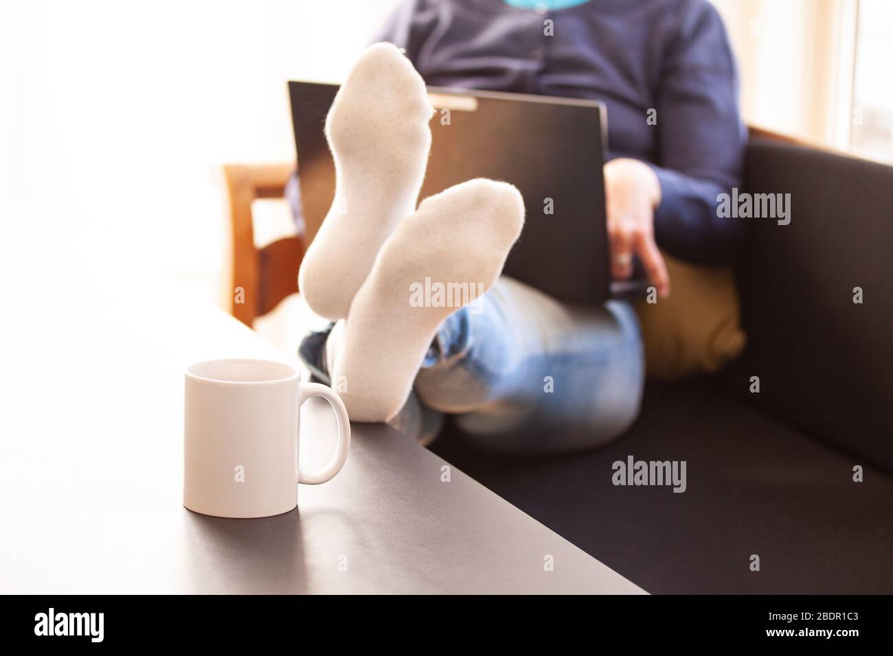 Donna seduta in un divano con un computer portatile e si trova a piedi su un tavolo dove è stata sistemata una tazza o una tazza. Lavorare da casa concetto. Messa a fuoco selettiva AN Foto Stock