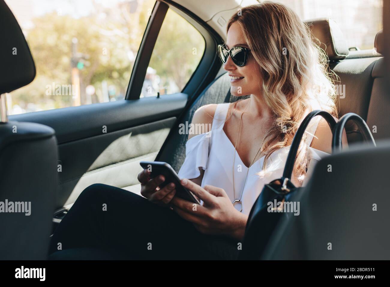 Agente immobiliare femminile in viaggio in auto. Donna d'affari che indossa occhiali da sole seduti sul sedile posteriore di un'auto che guarda fuori dalla finestra e sorridente. Foto Stock