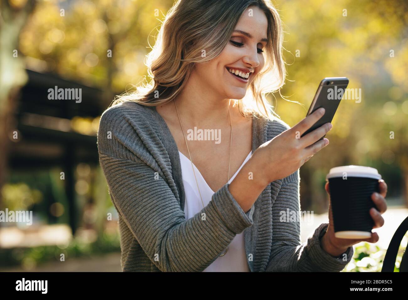 donna d'affari seduta all'aperto con il telefono. Agente immobiliare con una tazza di caffè in mano che legge un messaggio di testo sul suo cellulare fuori. Foto Stock