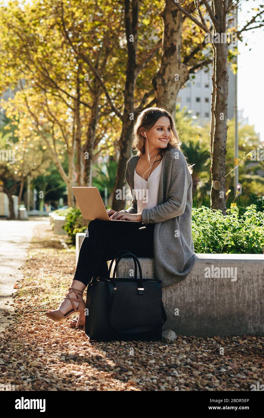 Giovane donna sorridente seduto all'aperto con il laptop. Donna che guarda via e sorridente mentre si siede fuori in città. Foto Stock