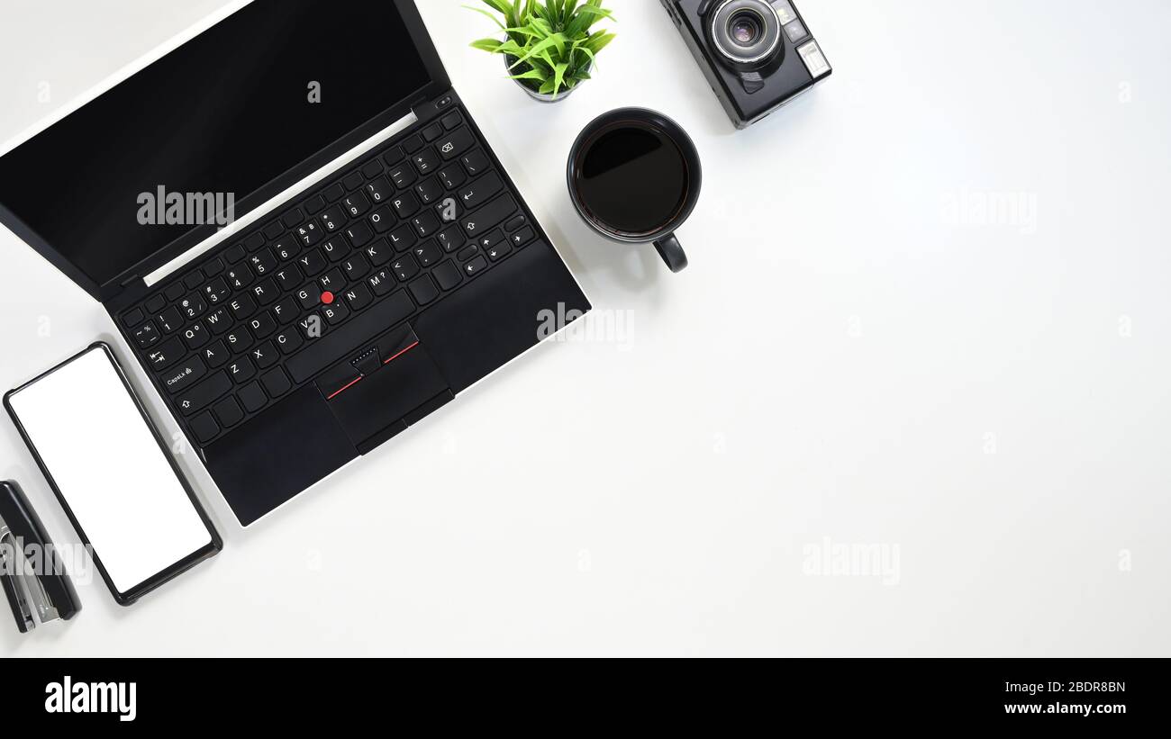 Immagine dall'alto del computer portatile nero che si mette su un tavolo bianco con tazza da caffè, fotocamera, pianta in vaso, smartphone bianco vuoto e cucitrice. Disposizione piatta Foto Stock