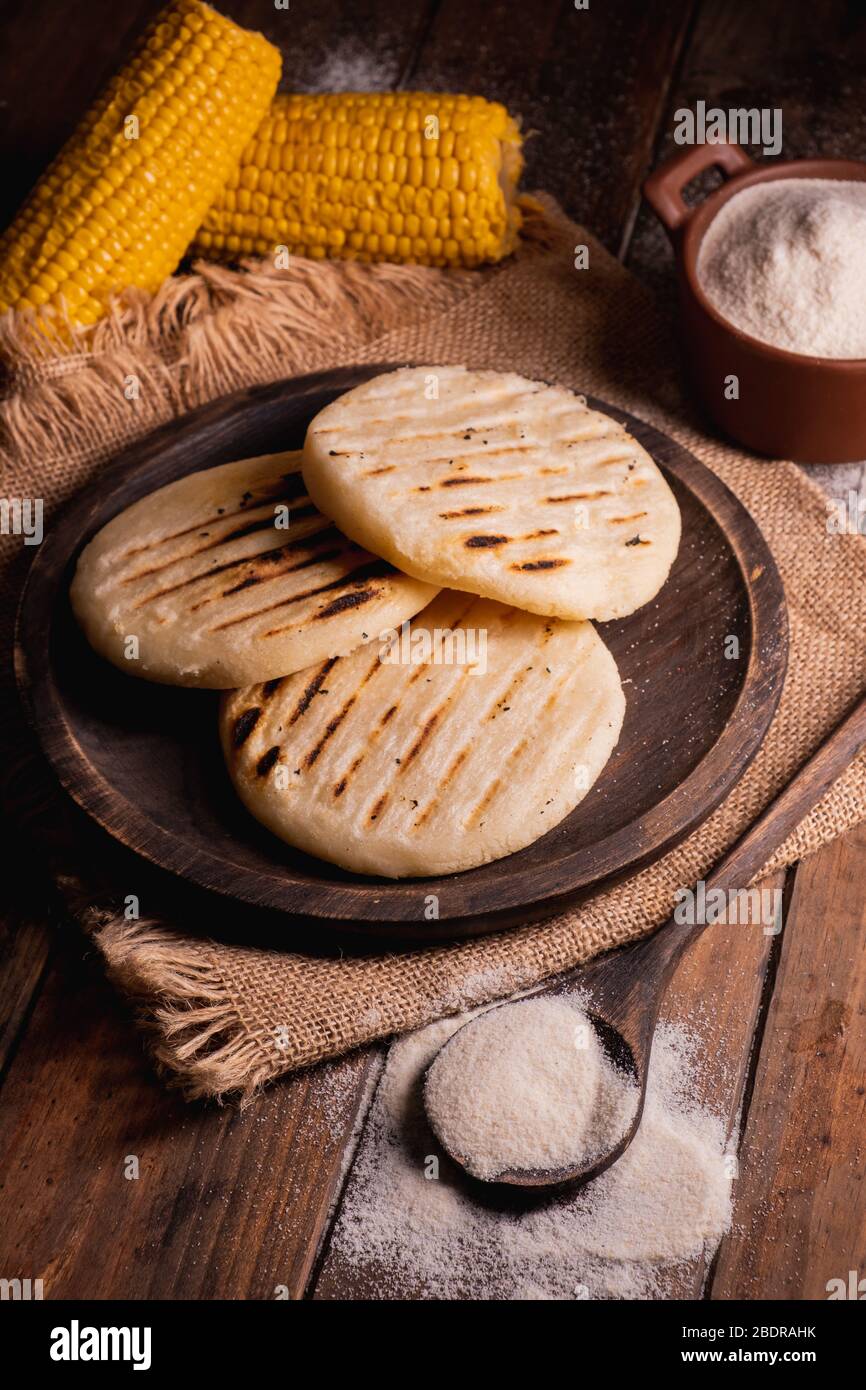 Arepas, cibo sudamericano a base di farina di mais. Tre arepas su un piatto  di legno e accanto ad esso un cucchiaio e una ciotola piena di farina di  mais su un