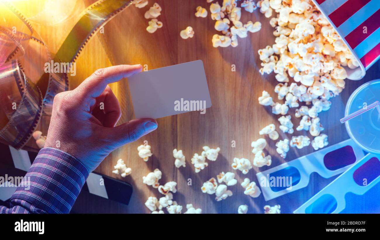 Mano che tiene un cinema vuoto promo carta o un biglietto; popcorn, filmina e battaglio sullo sfondo; filmati e concetto di intrattenimento Foto Stock