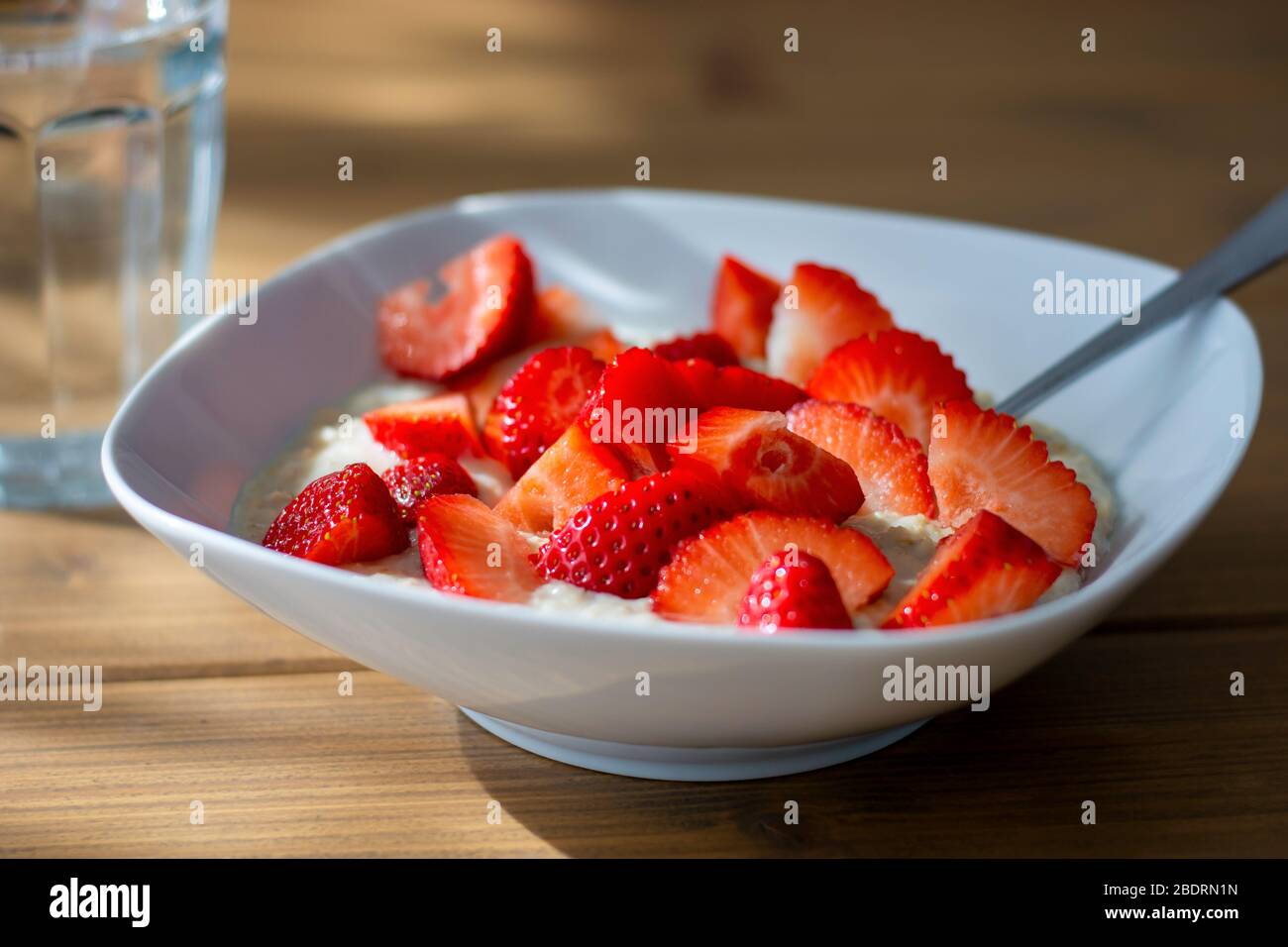 Ciotola di porridge d'avena fatta in casa con fragole a fette e un bicchiere d'acqua sul tavolo alla luce del sole del mattino. Pasto sano. Porridge avena Foto Stock