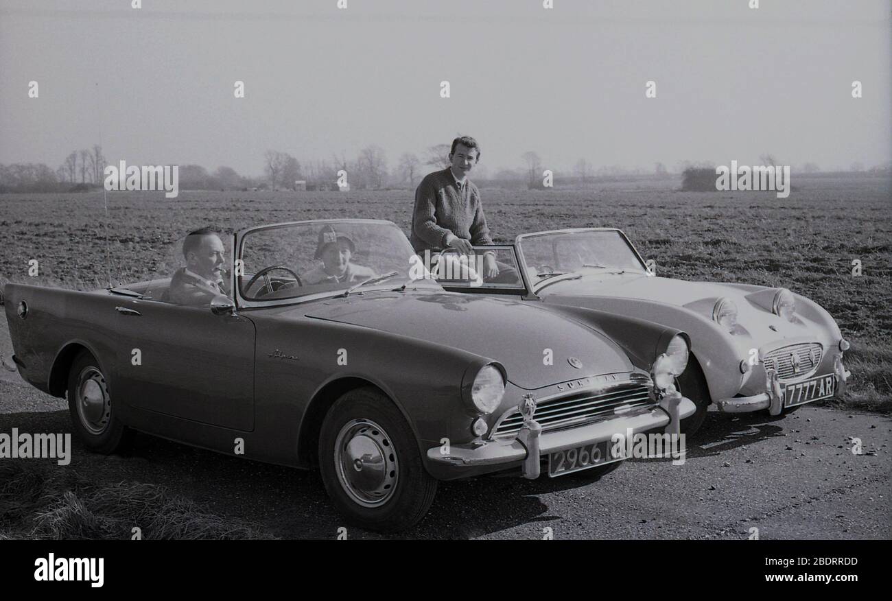 Anni '60, storico, automobilistico, due auto sportive open-top 2 porte dell'epoca parcheggiate l'una accanto all'altra accanto a un campo di colture, vicino a Eastholm Green, Letchworth Garden City, Herts, Inghilterra, Regno Unito. Una coppia matura è seduta nella loro Sunbeam Alpine, con il Signore che fuma un tubo. Un uomo più giovane, forse il figlio, è seduto in un Austin Healey Sprite MK1, noto come 'Frogeye' a causa dell'aspetto dei fari delle auto sul cofano. Foto Stock