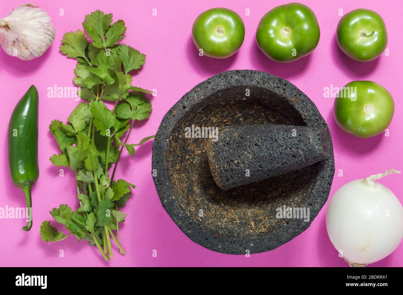 Salsa verde cruda ingredienti, cibo messicano. Tomatillos, cipolla, aglio, peperoncino e coriandolo schiacciati in una molcajete. Ingredienti verdi su rosa Foto Stock
