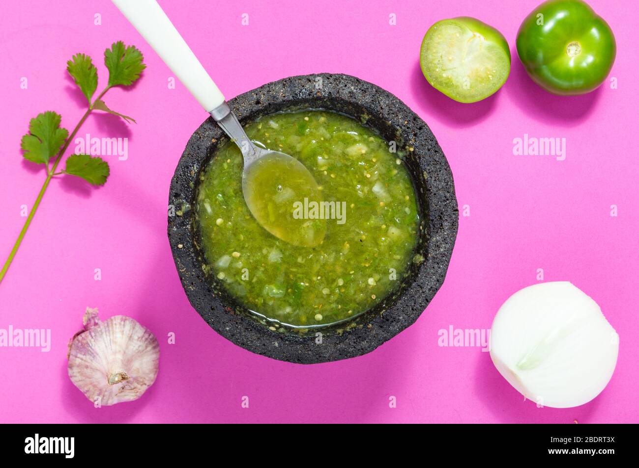 Salsa verde cruda ingredienti, cibo messicano. Tomatillos, cipolla, aglio, peperoncino e coriandolo schiacciati in una molcajete. Ingredienti verdi su rosa Foto Stock