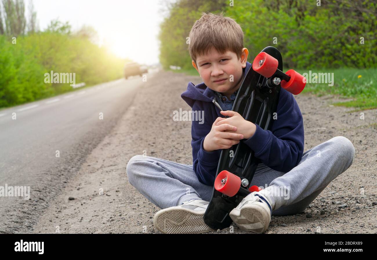 Un ragazzo in abiti sportivi con un pattino che riposa vicino alla strada all'alba. Foto Stock