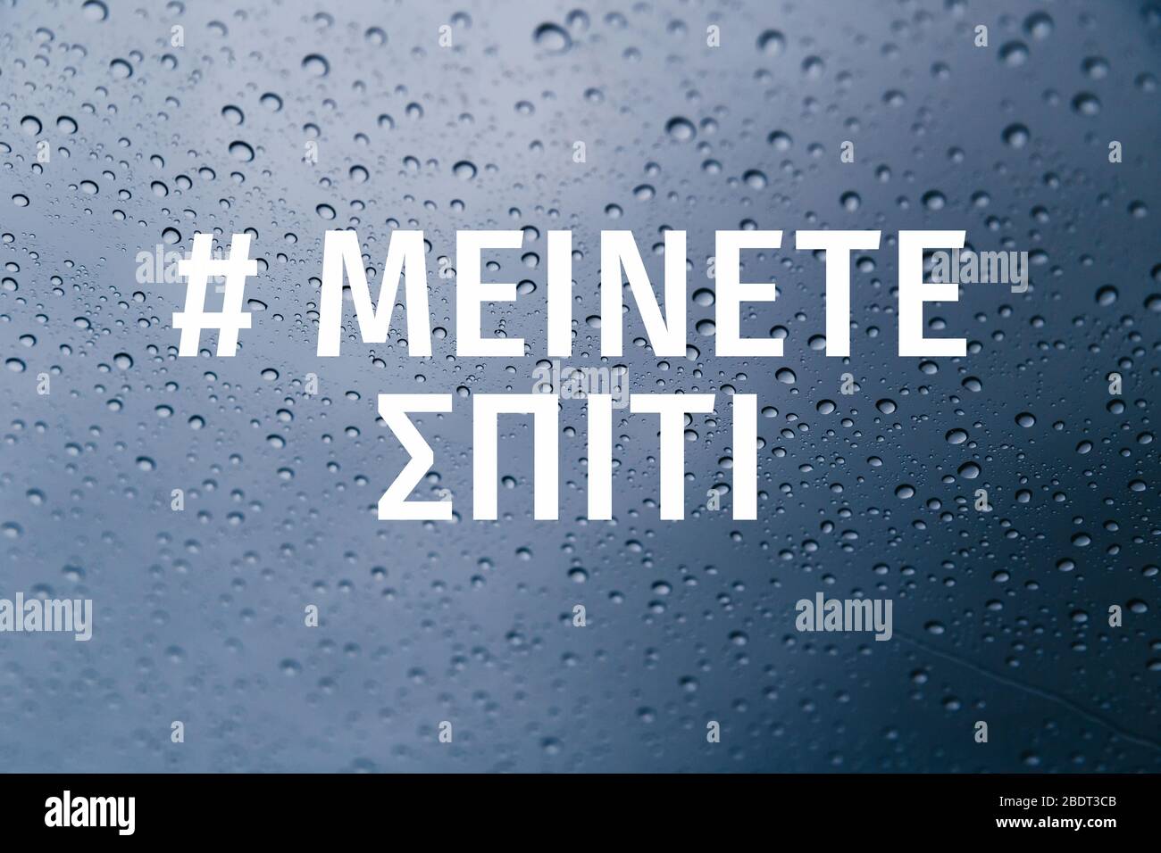 Rimani a casa nella campagna dei social media in lingua greca per la prevenzione del coronavirus. Soggiorno a casa concetto di sicurezza. Primo piano di gocce di pioggia sul finestrino Foto Stock