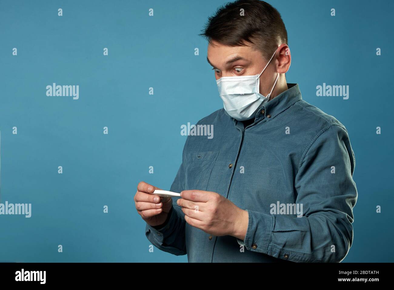 Giovane uomo in tessuto casual indossare maschera medica protettiva eccitato, stressato e spaventato dopo la lettura molto alta temperatura risultato da digitale Foto Stock