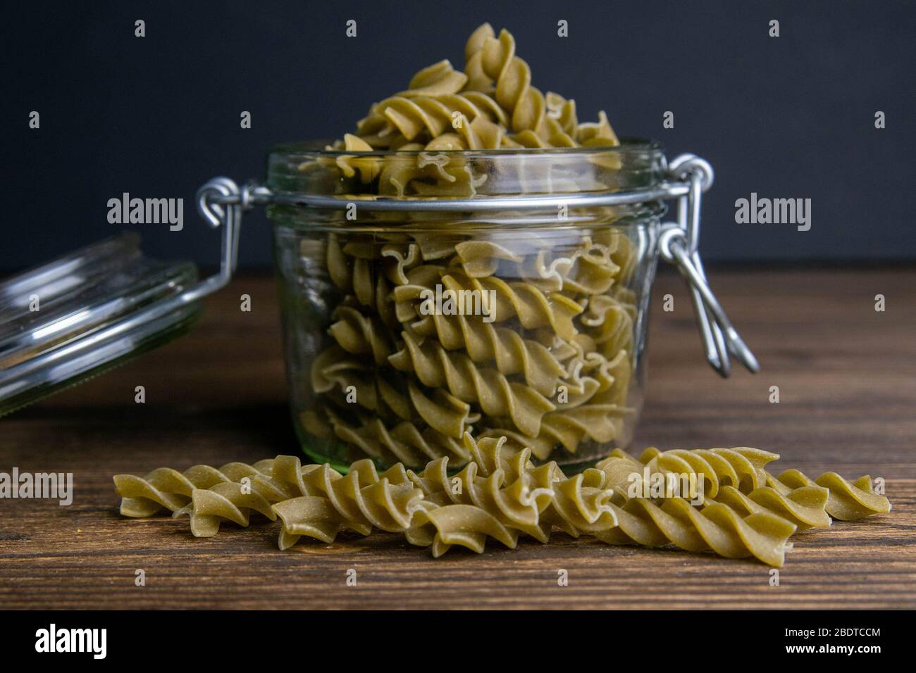 Vaso di vetro riempito con pasta secca spirale fusilli e sparso intorno su sfondo di legno scuro Foto Stock