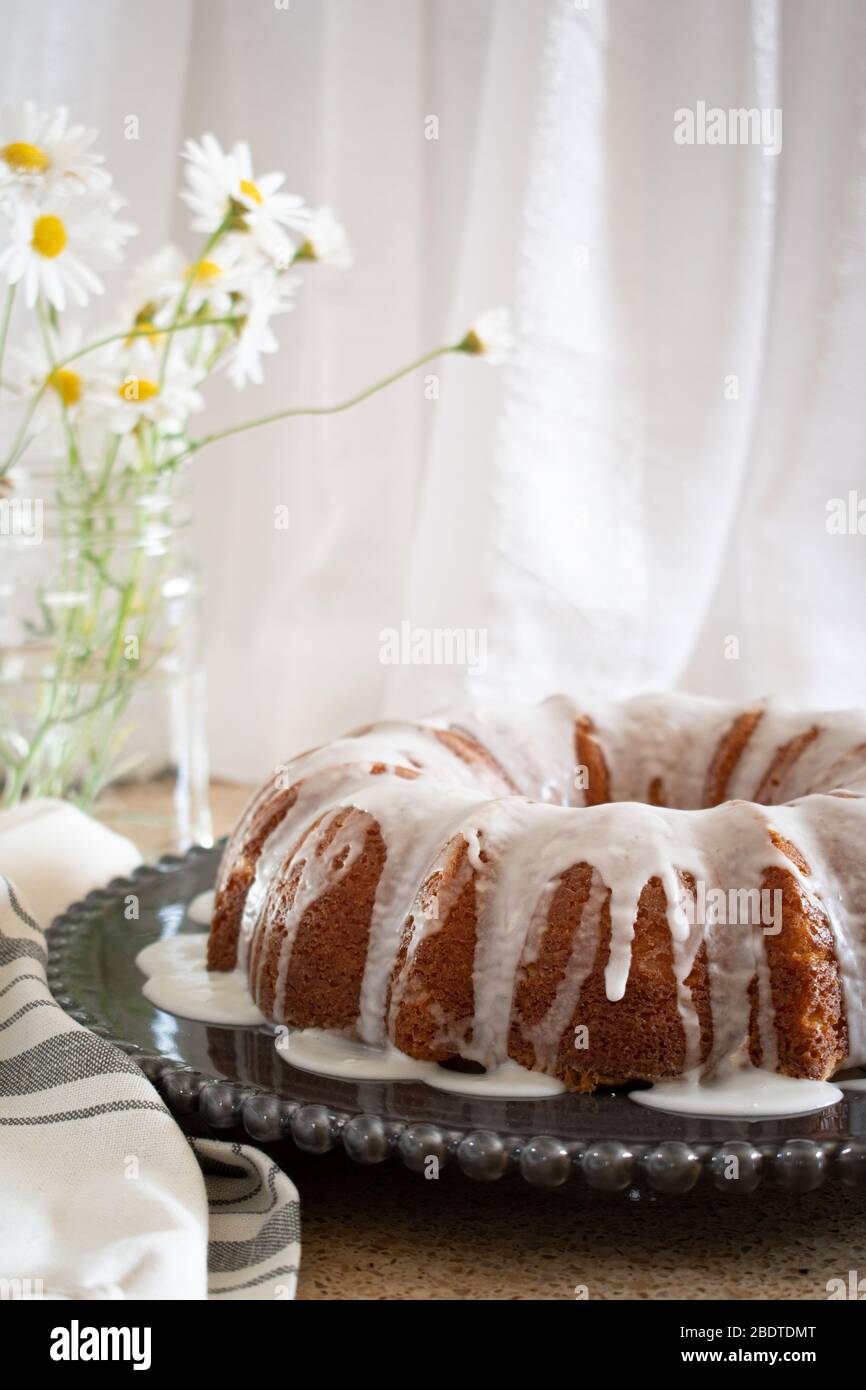 Torta al limone con fiocco di ricotta e glassa, esposta su un piatto decorativo nero con fiori Daisy in un vaso sullo sfondo. Foto Stock