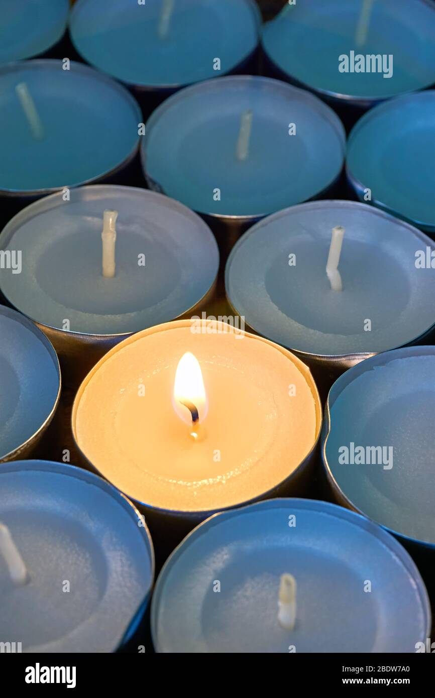 Primo piano delle luci del tè con una accesa. Una singola fiamma calda arancione tra candele in ombra blu. Natale, lutto o condoglienza concetto. Foto Stock