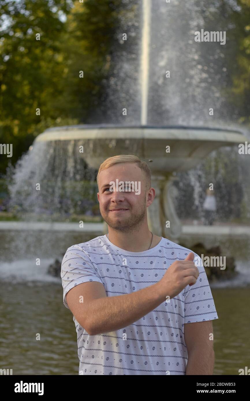 un giovane biondo in una maglietta leggera lancia una moneta nella fontana per una buona fortuna Foto Stock