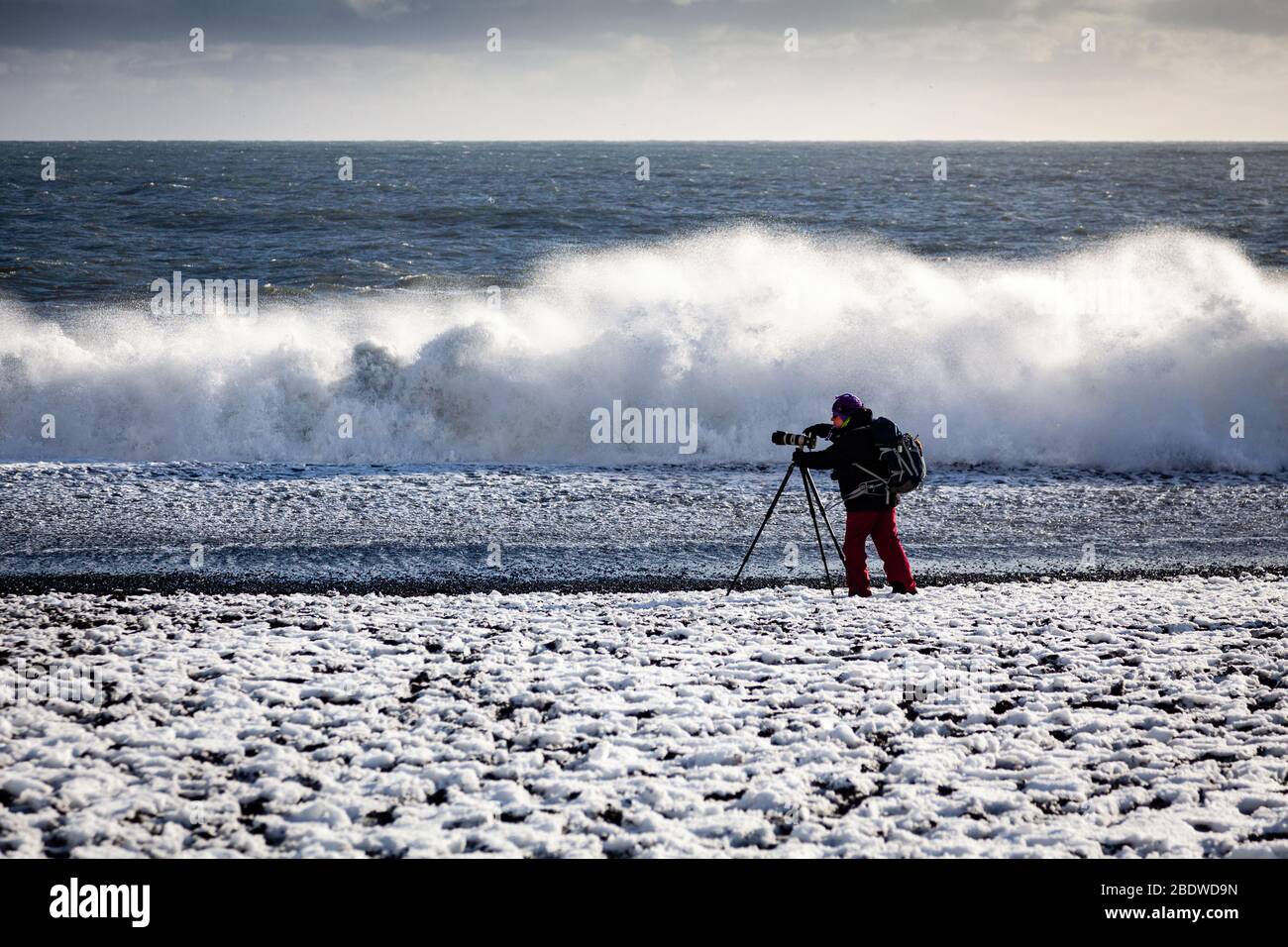 Un fotografo che fotografa le onde delle sneaker che si infrangono sulla spiaggia di sabbia nera di Reynisfjara coperta di neve a Vík í Mýrdal, sulla costa meridionale dell'Islanda Foto Stock