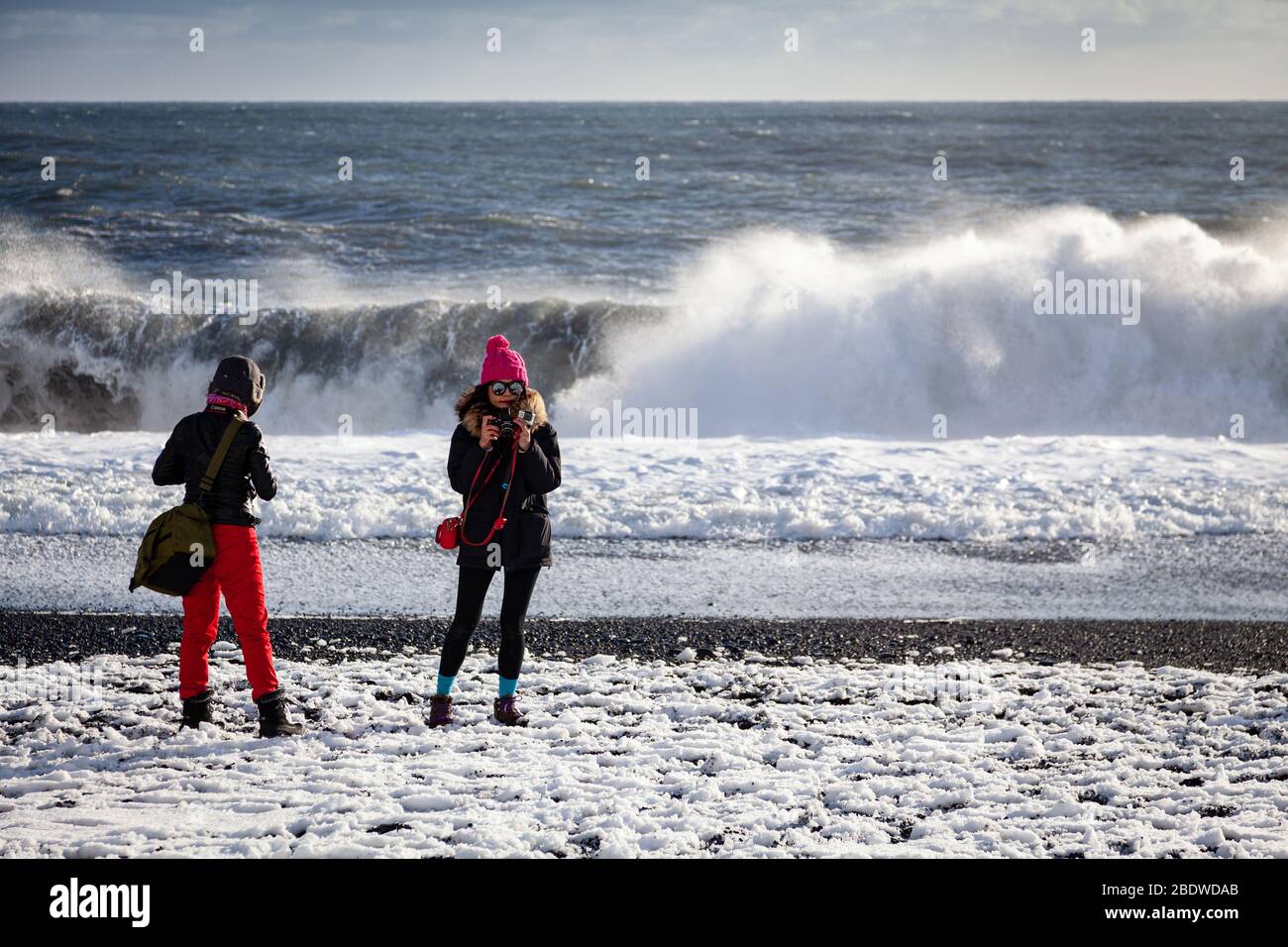Turisti in abiti invernali scattare foto di onde sneaker rompere su Reynisfjara spiaggia di sabbia nera coperta di neve vicino a Vík í Mýrdal in Islanda Foto Stock