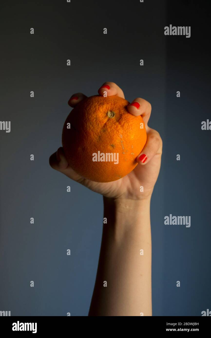 Mano femminile con chiodi dipinti di rosso che reggono un arancione su uno sfondo grigio. Foto Stock