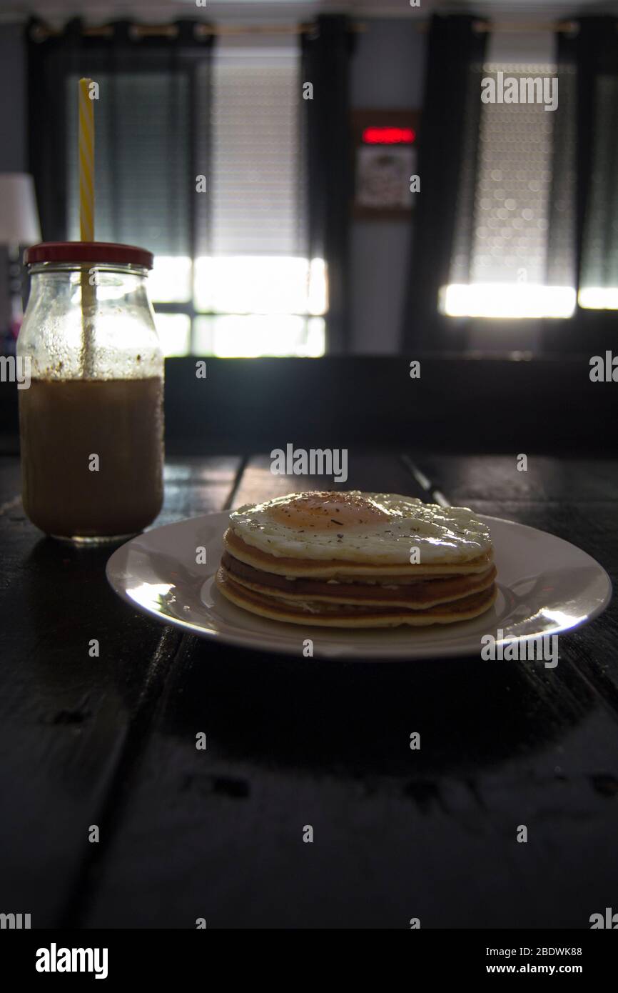 Pancake con uova e caffè in un vaso con paglia gialla su tavolo di legno nero e con sfondo non focalizzato. Foto Stock