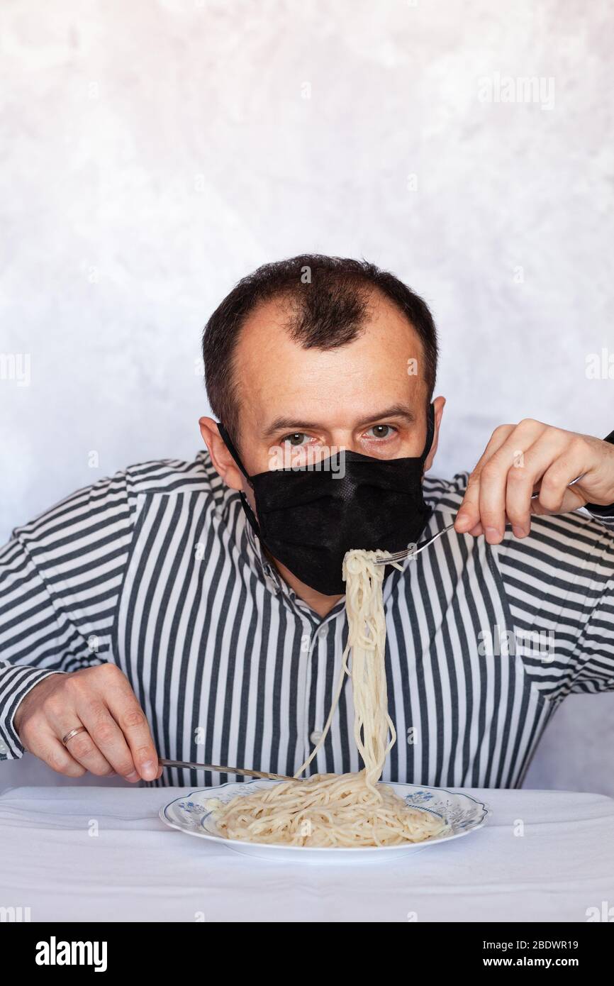 Un bell'uomo in una maschera medica nera mangia la pasta sul tavolo contro lo sfondo chiaro Foto Stock