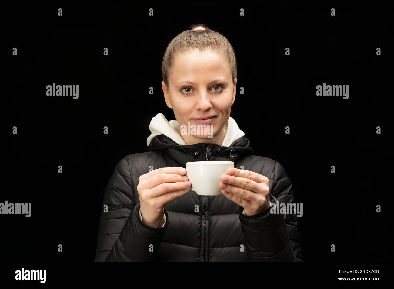 Una giovane ragazza europea beve tè o caffè da una tazza bianca e sorride. In una giacca nera calda su uno sfondo scuro Foto Stock