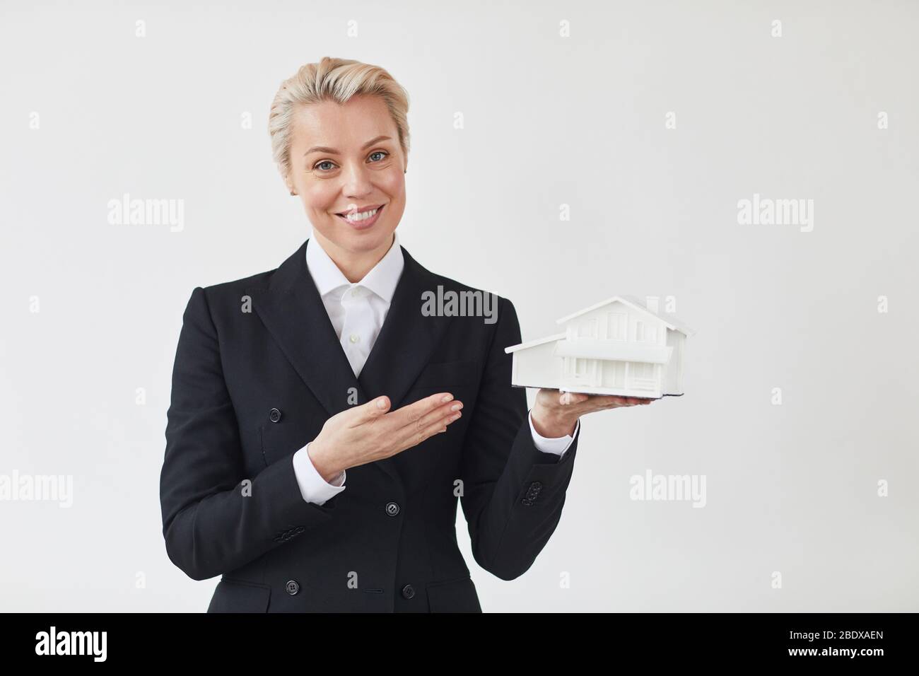Maturo realtor in abito nero che tiene modello della casa e sorridente a macchina fotografica isolato su sfondo bianco Foto Stock