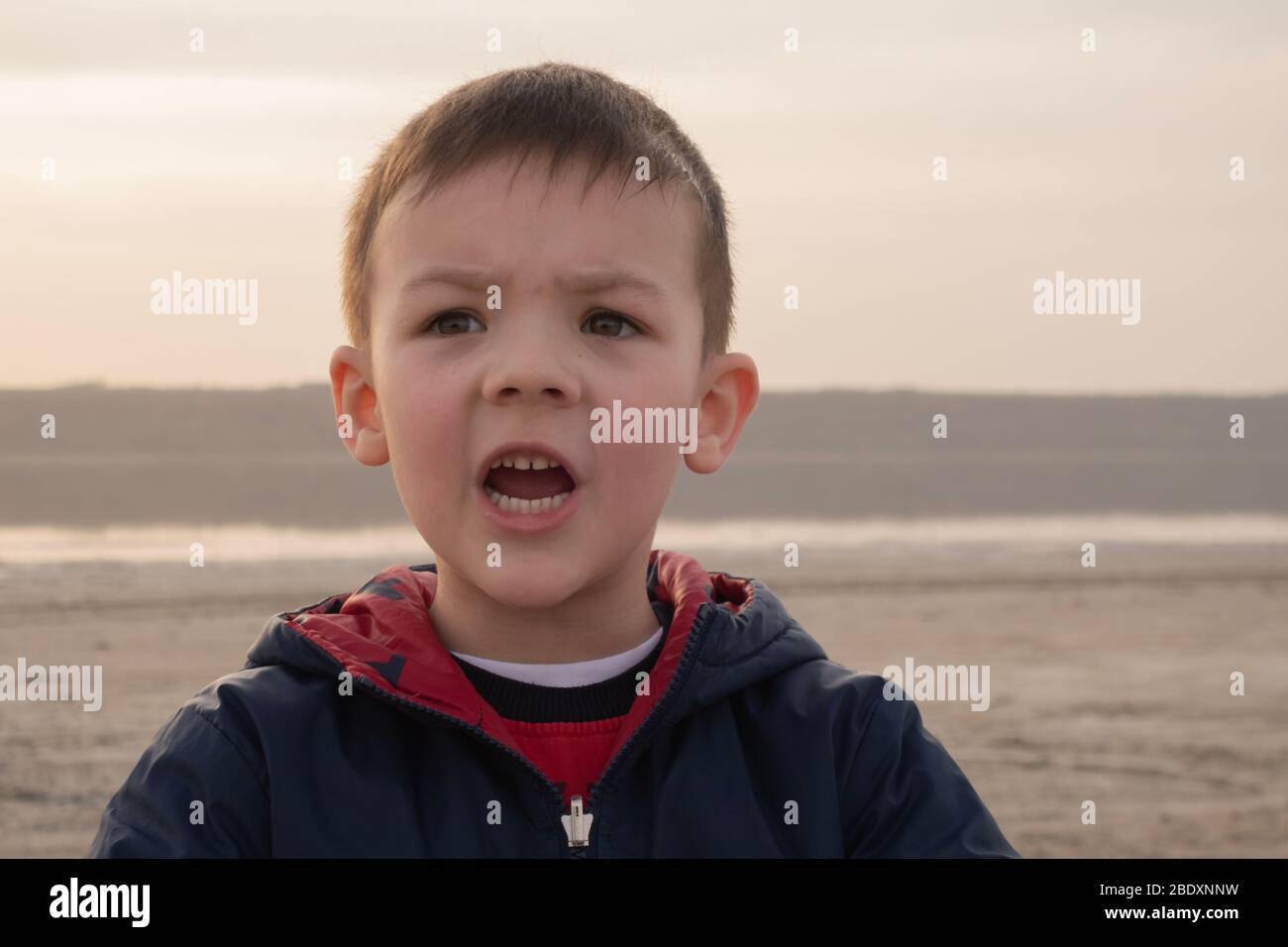 Un ragazzo di 4 anni in una giacca blu su una spiaggia vuota. Il bambino urla e si innervosisce. Foto Stock