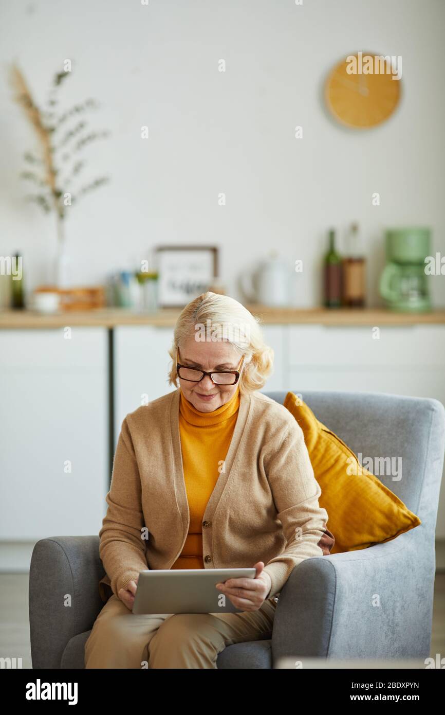 Donna matura in occhiali seduti su poltrona e con tablet digitale in salotto a casa Foto Stock