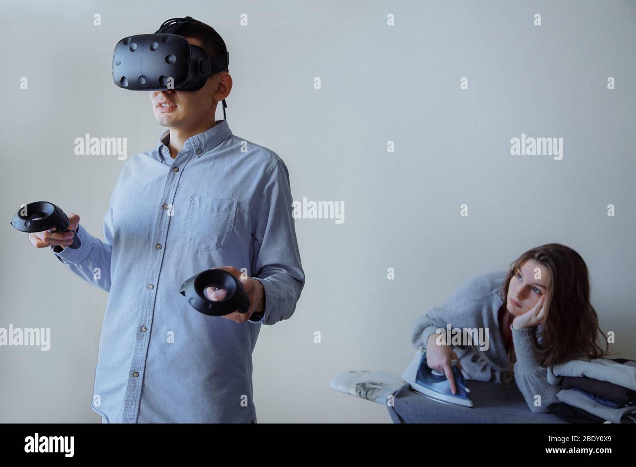 Un uomo con entusiasmo gioca ai videogiochi in realtà virtuale. La moglie sta facendo i lavori domestici in questo momento. Foto Stock