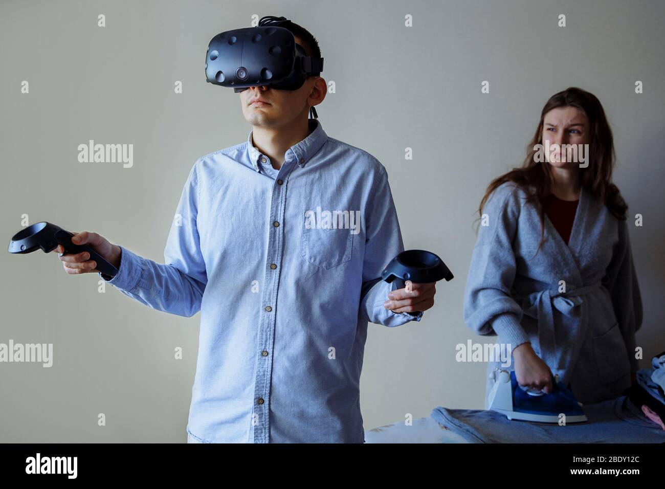 Un uomo con una camicia blu in denim gioca in realtà virtuale. Moglie che strofinava le camicie e gli sguardi offesi al marito Foto Stock