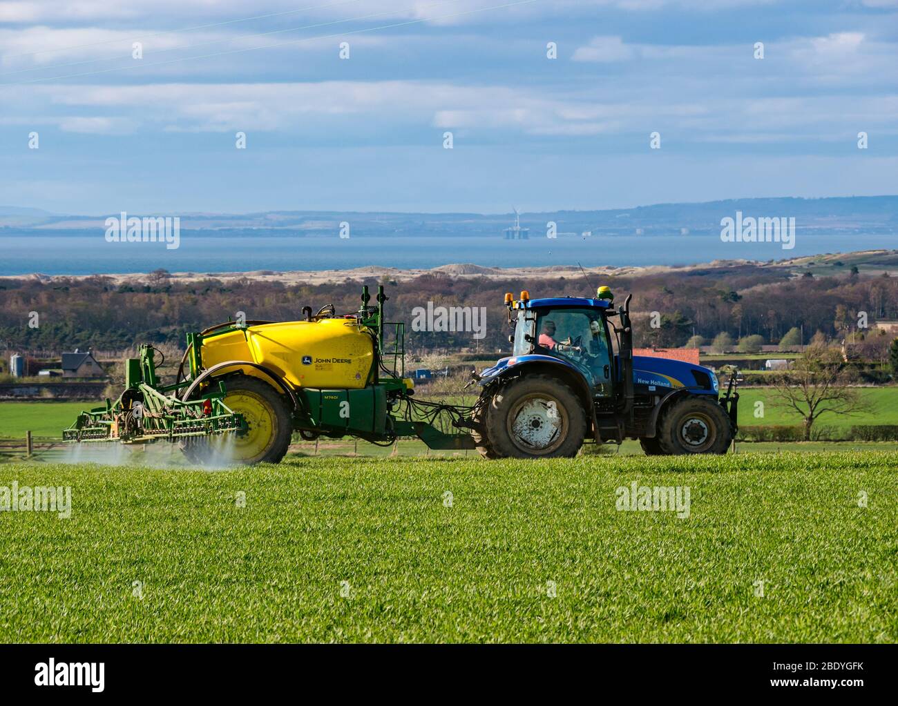Trattore che irrora il campo di coltura in un paesaggio arabile con vista di Firth of Forth, East Lothian, Scozia, Regno Unito Foto Stock