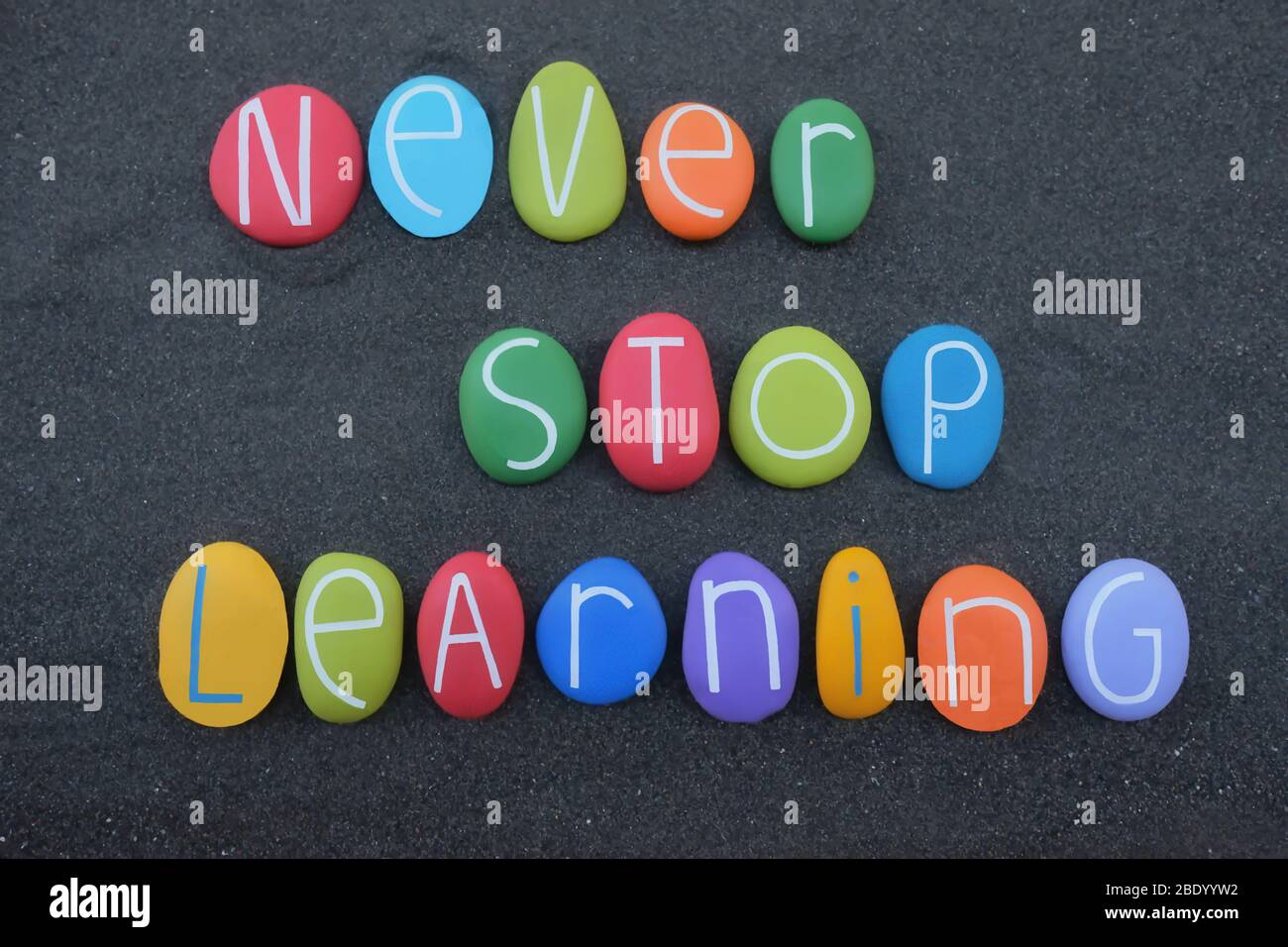 Never Stop Learning, slogan motivazionale composto da lettere in pietra multicolore su sabbia vulcanica nera Foto Stock