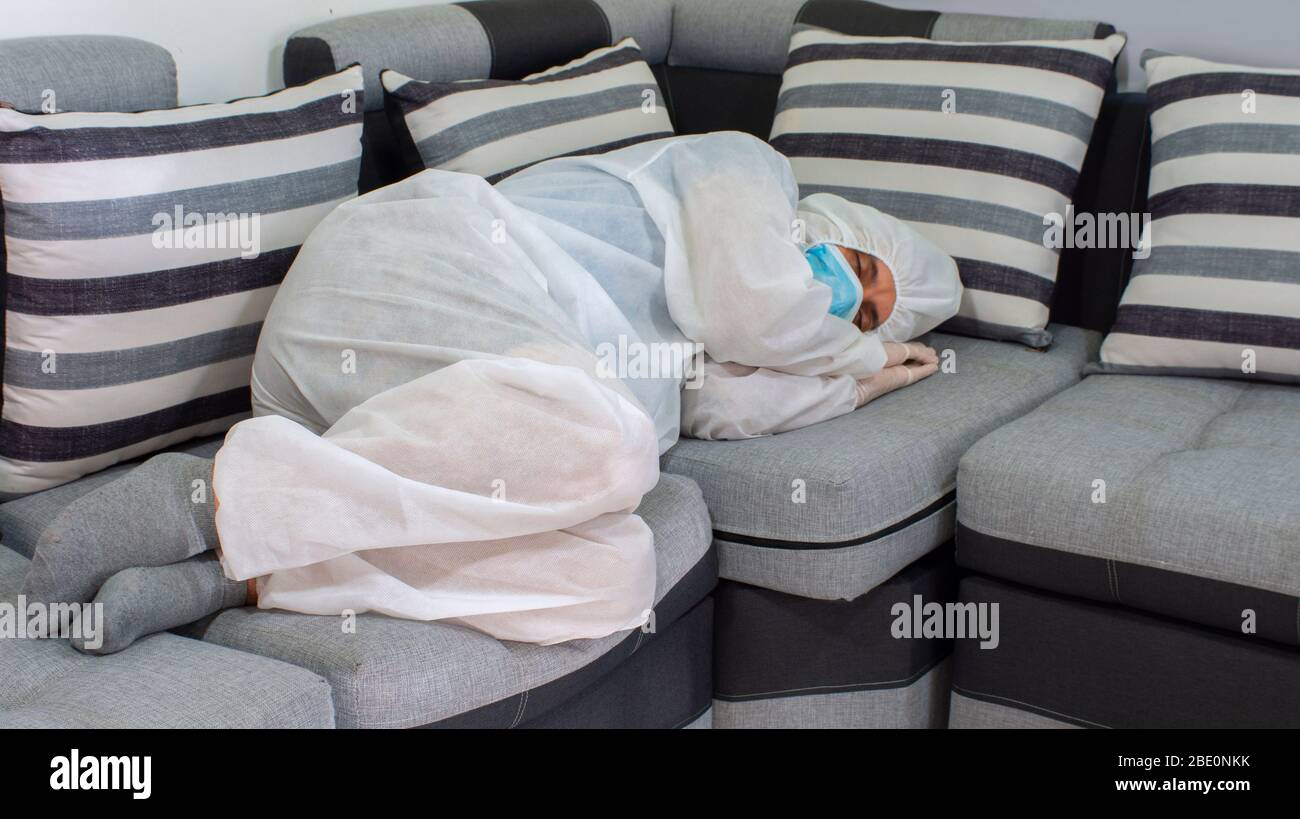 Vista frontale del medico con maschera blu e tuta bioprotettiva che dorme su un grande divano in tonalità grigie su sfondo bianco Foto Stock