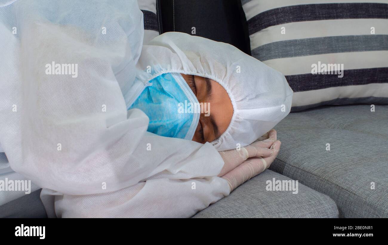 Vista ravvicinata del medico con maschera blu e tuta bioprotettiva che dorme su un grande divano in tonalità grigie di notte Foto Stock