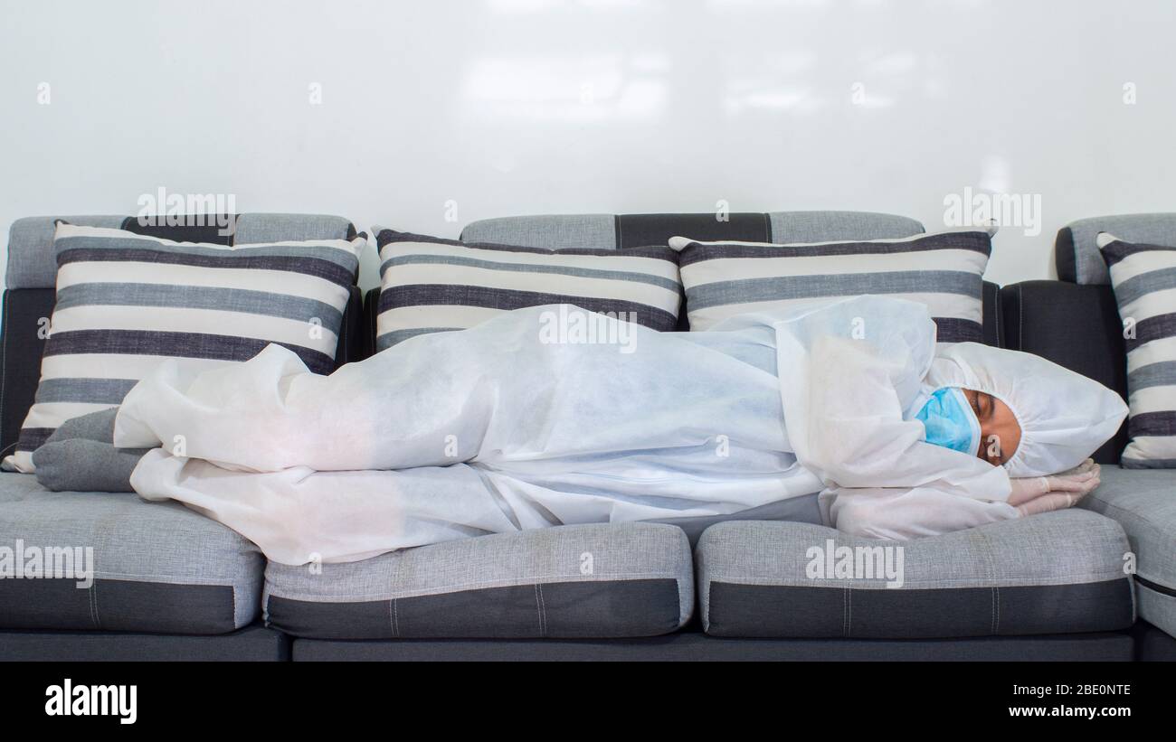 Vista frontale del medico con maschera blu e tuta bioprotettiva che dorme su un grande divano in tonalità grigie di notte su sfondo bianco Foto Stock