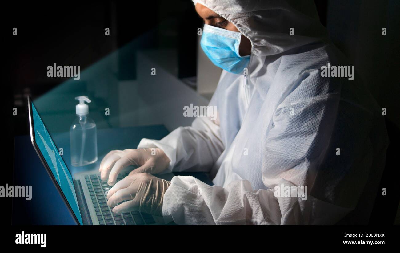 Vista laterale del medico con maschera blu e tuta bioprotettiva concentrata in ufficio con il suo laptop sulla sua scrivania con bottiglia di gel di alcol w Foto Stock
