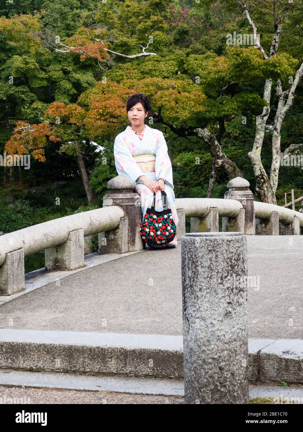 Kyoto, Giappone - 30 settembre 2015: Giovane ragazza in kimono bello seduto su un ponte di pietra in giardino giapponese tradizionale Foto Stock