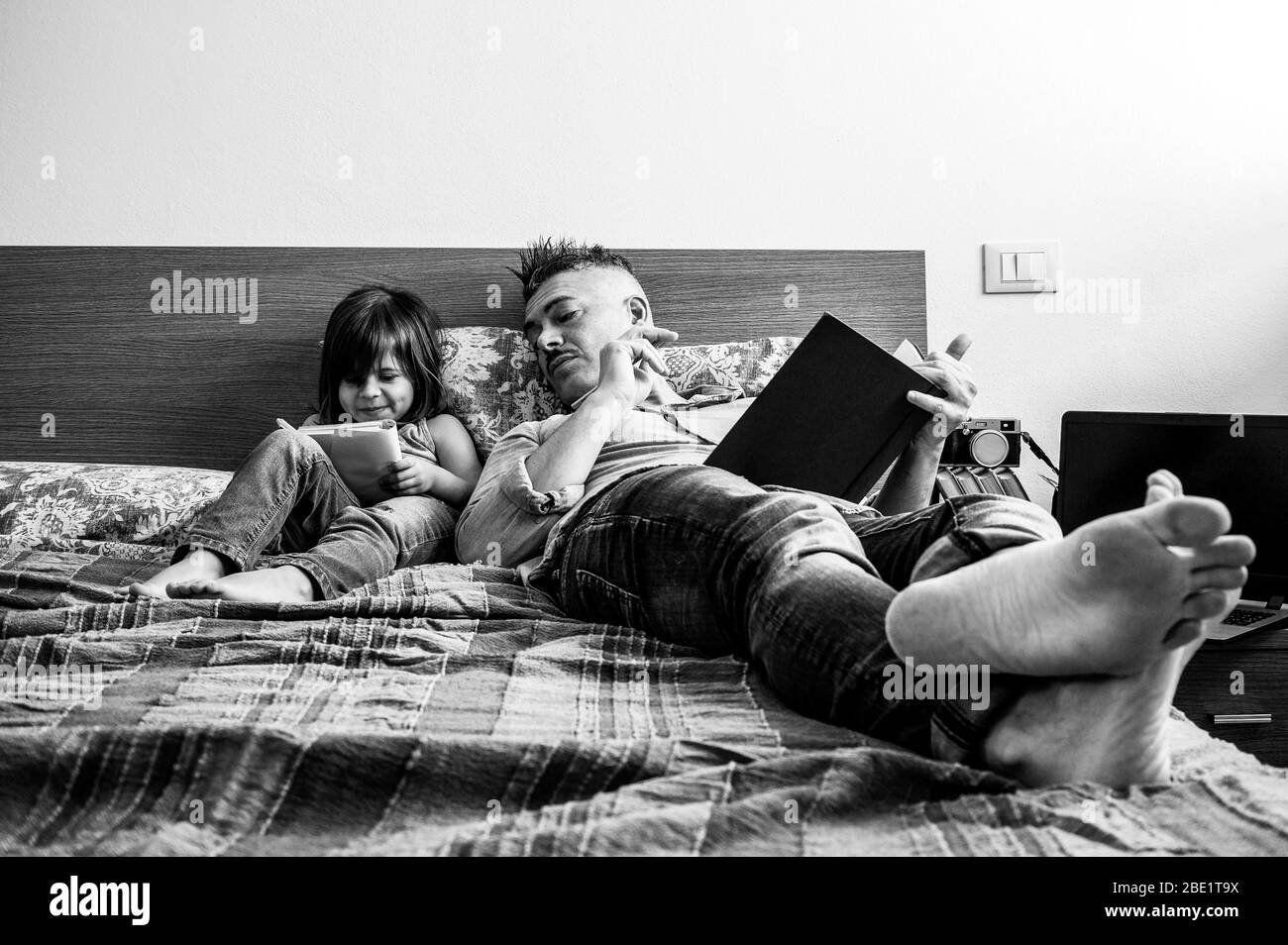 Padre e figlia in abiti denim casual sono seduti a letto insieme. La ragazza che ha divertimento di scrittura o disegno mentre il padre la guarda. Infanzia Foto Stock