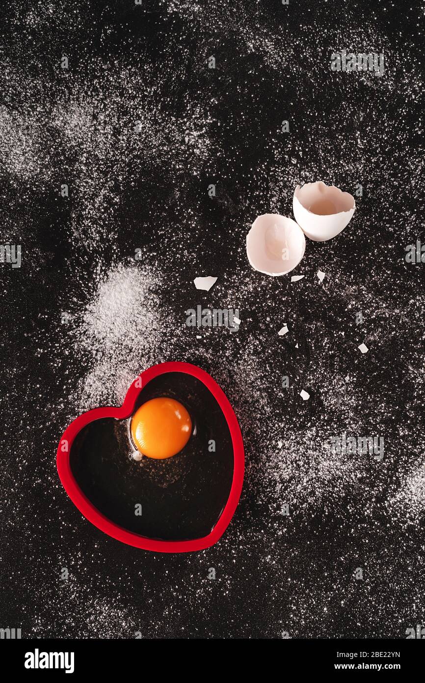 Immagine chiave bassa di un bianco crudo uova biologiche su sfondo nero cosparso di farina. Un uovo rotto aperto sulla forma rossa a forma di cuore di silicone. Cucina Foto Stock