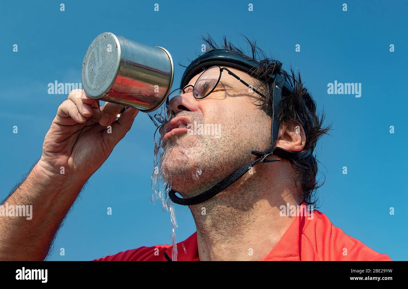 Ritratto di un ciclista stanco in casco retrò beve acqua da tazza, versione con filtro. Bevanda dissetata di biker contro il cielo blu. Foto Stock