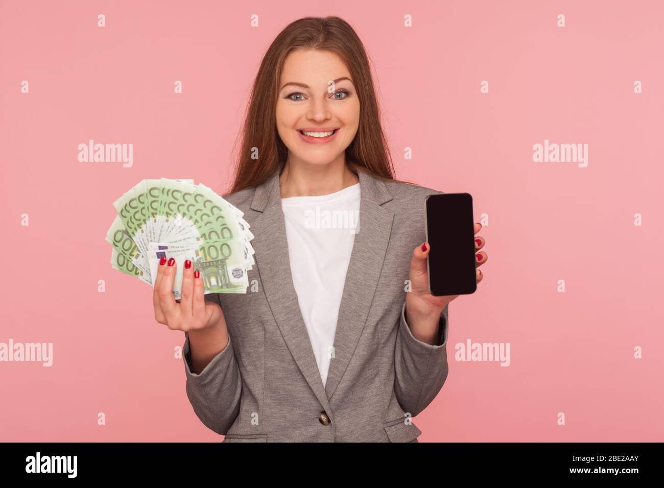 Domanda di prestito, richiedere online. Ritratto di felice donna d'affari sorridente in giacca in abito con banconote in euro e cellulare, soddisfatto con il divieto di telefonia mobile Foto Stock