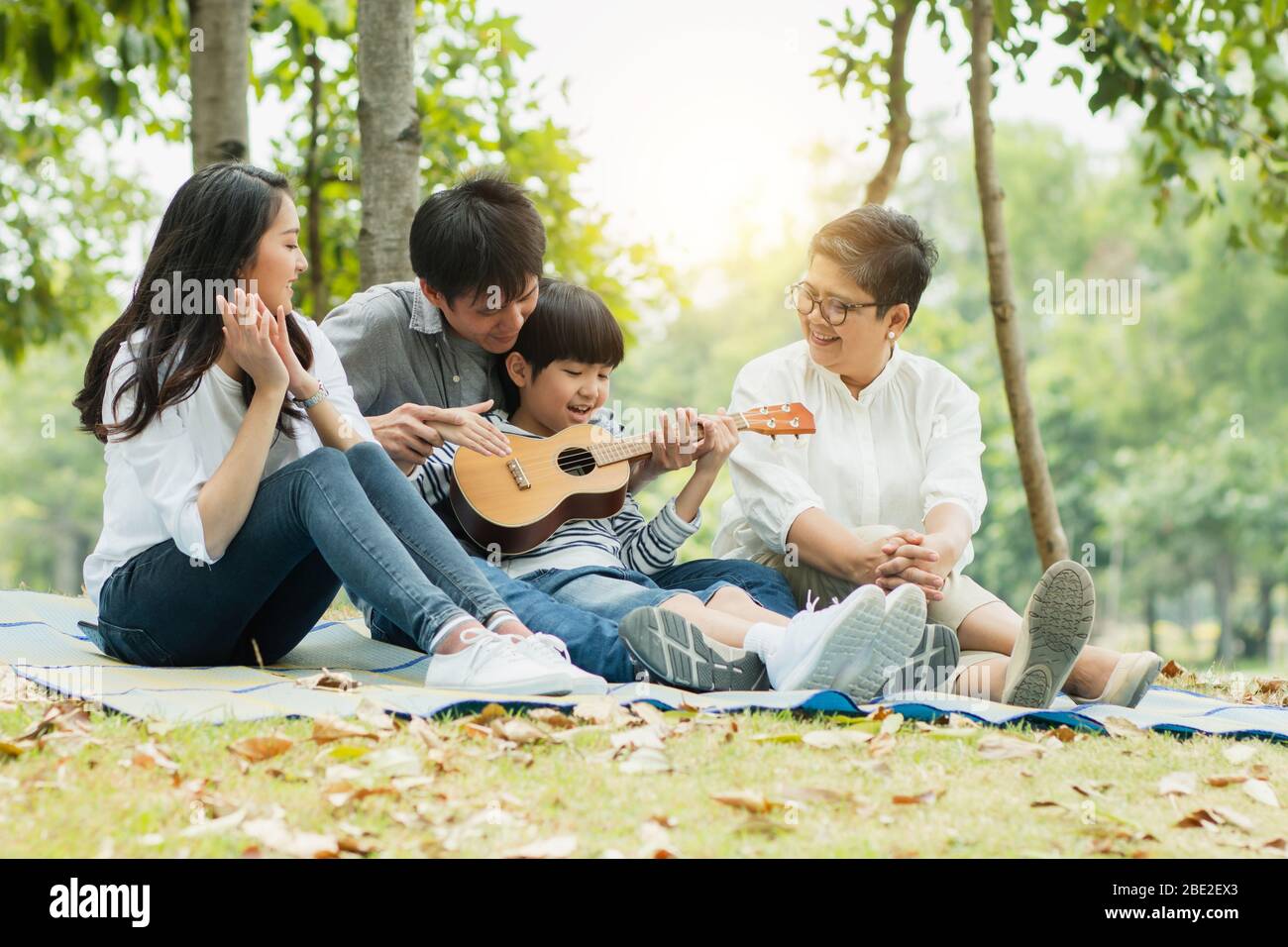 Famiglia felice con nonna, mamma con papà che insegnano figlio a suonare la chitarra e cantare una canzone nel parco, godere e rilassarsi persone pic-nic all'aperto Foto Stock