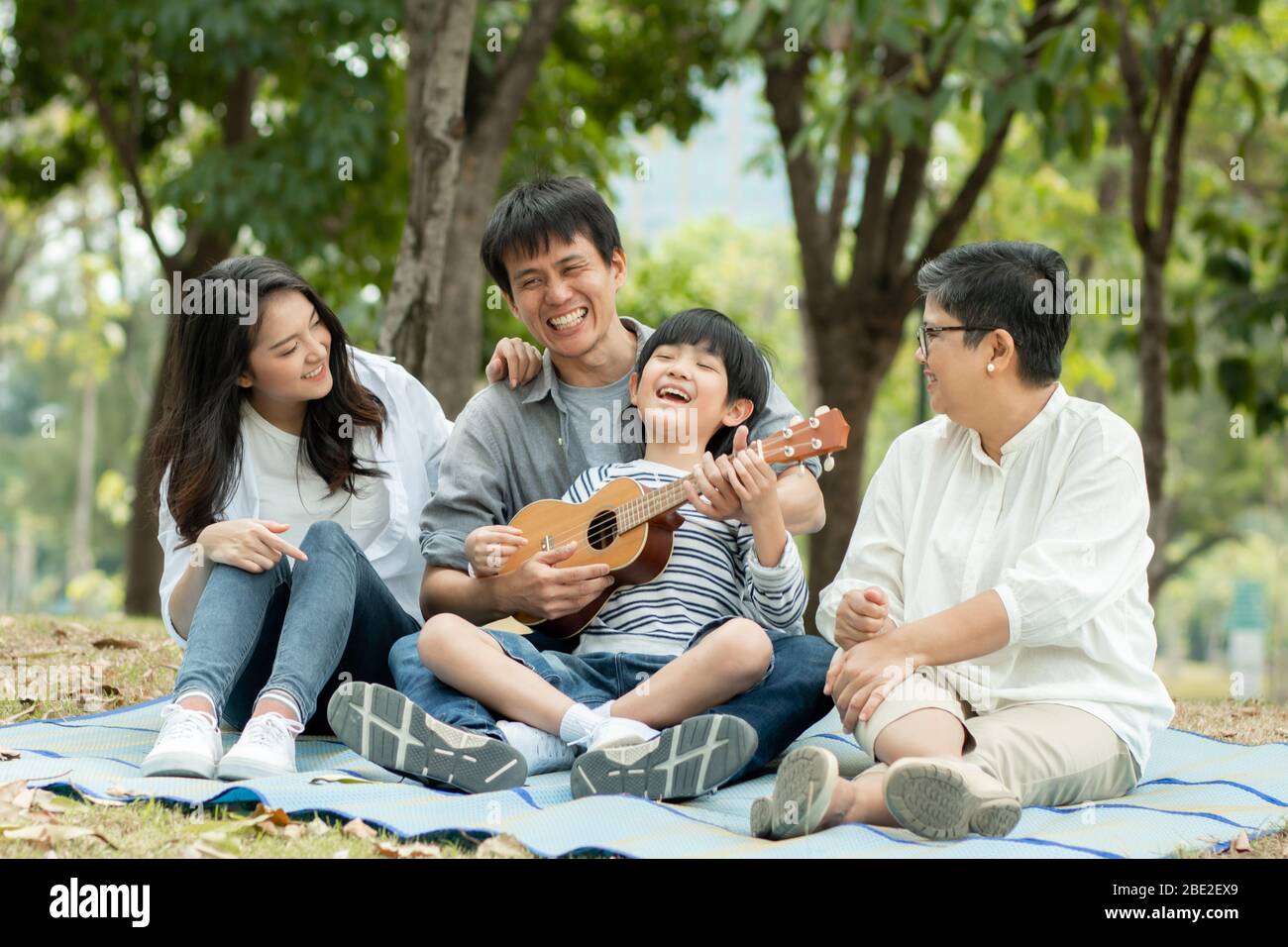 Famiglia felice con nonna, mamma con papà che insegnano figlio a suonare la chitarra e cantare una canzone nel parco, godere e rilassarsi persone pic-nic all'aperto Foto Stock