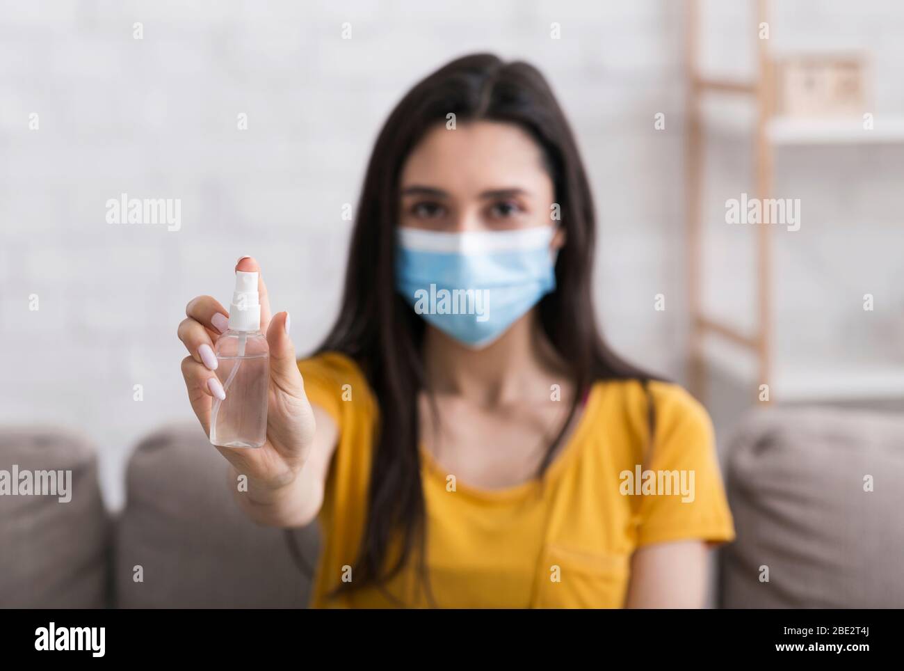 Prevenzione COVID-19. Ragazza in maschera protettiva che tiene bottiglia di igienizzatore a casa, fuoco selettivo Foto Stock