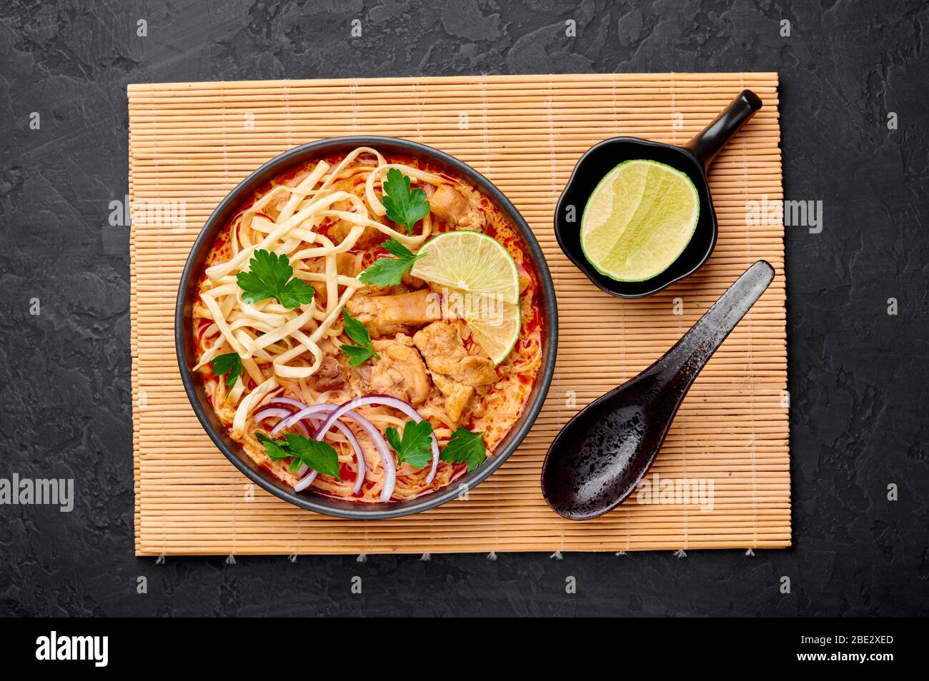 Khao Soi Gai o zuppa di nodle di curry di cocco tailandese in una ciotola nera su sfondo di ardesia scuro. Khao Soy è piatto di cucina thailandese con tagliatelle, carne di pollo, r Foto Stock