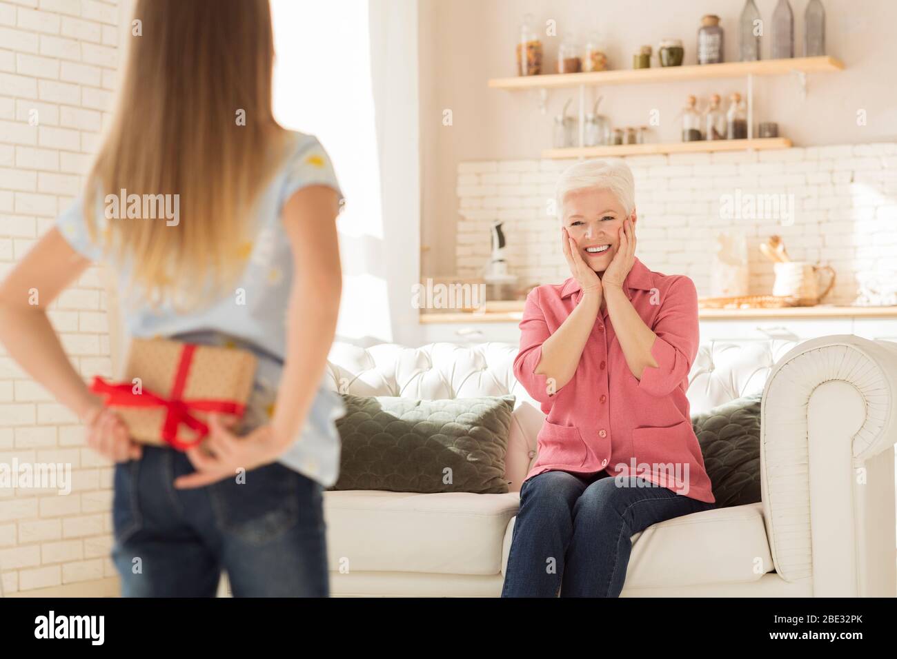 Bambina con regalo dietro la schiena e nonna emozionata in soggiorno Foto Stock