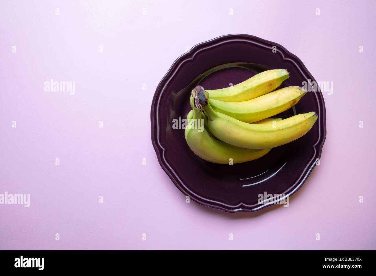 un'elegante immagine color pastello di un mazzo di banane, un piatto viola decorativo con sfondo rosa, piatto, vista dall'alto Foto Stock