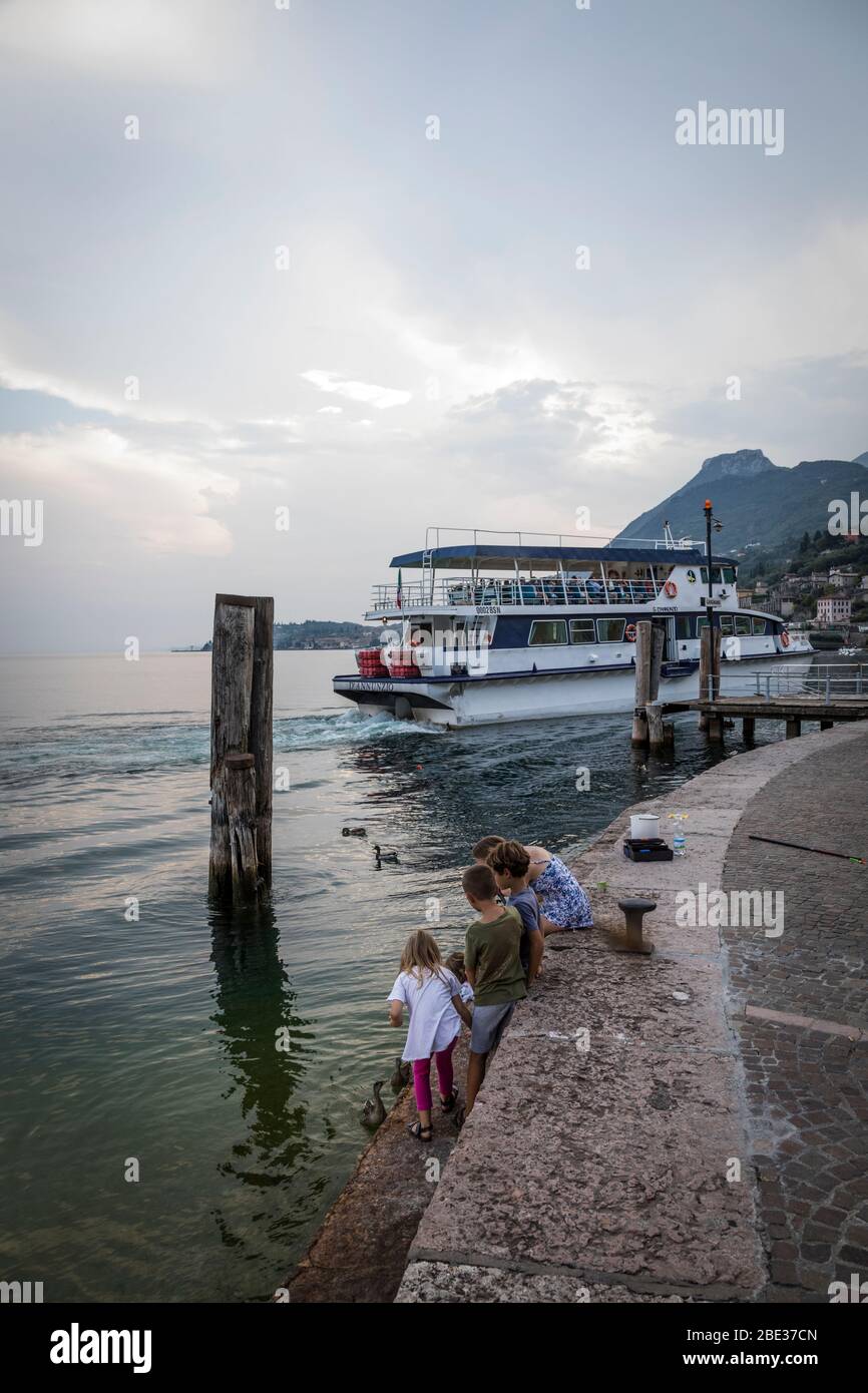 Un traghetto si attracca sullo sfondo mentre i bambini nutrono le anatre sulla riva del Lago di Garda a Gargnano, Italia. Foto Stock