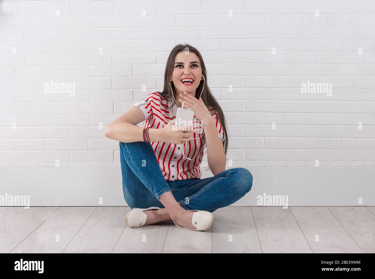 Ridendo giovane donna felice con telefono cellulare e auricolari, seduta a gambe incrociate, guardando su isolato bianco muro sfondo di mattoni. Una persona singola. Foto Stock