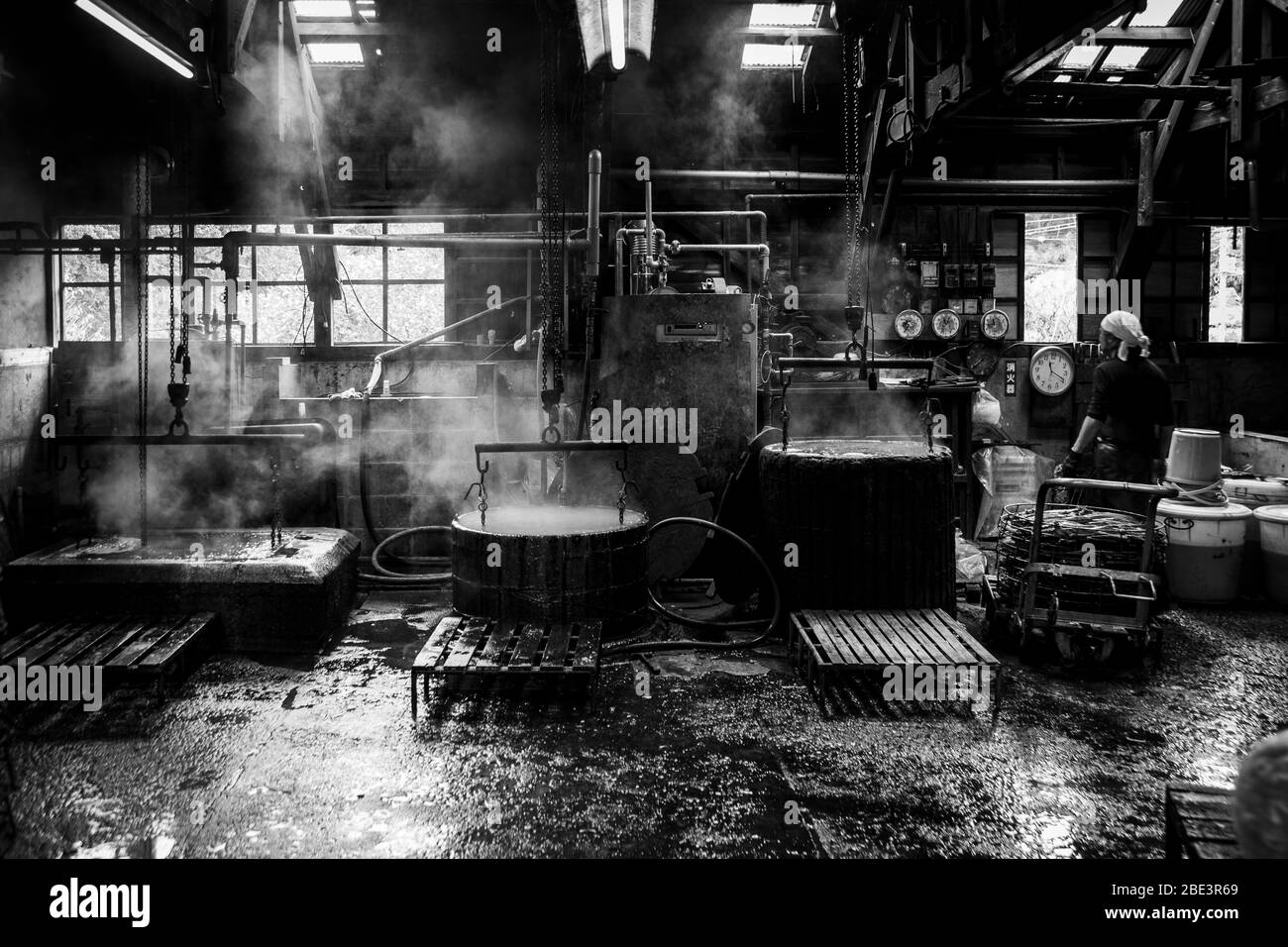 Un bac di legno pieno d'acqua e fumante si erge tra tubi metallici e catene nell'officina della manifattura Kanesa katsuobushi. In fondo a destra, un operaio si trova vicino al muro con un orologio e quadri elettrici. Nishiizu-Cho, Prefettura di Shizuoka, Giappone. Foto Stock