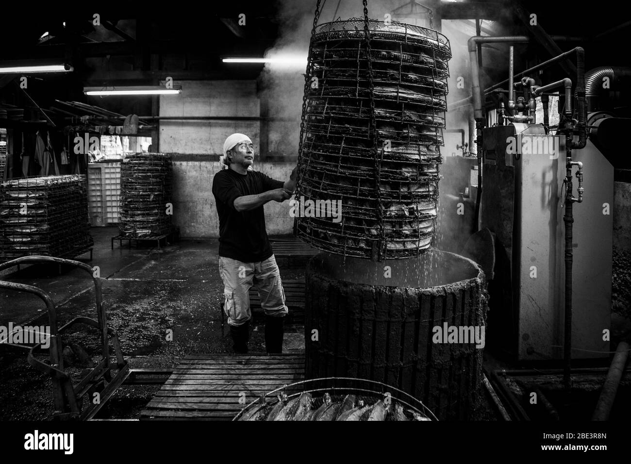 Yasuhisa Serizawa continua a sollevare la torre di cestini fuori dalla vasca, cestini di filo vuoti impilati sullo sfondo accanto alle macchine della fabbrica Kanesa katsuobushi. Nishiizu-Cho, Prefettura di Shizuoka, Giappone. Foto Stock